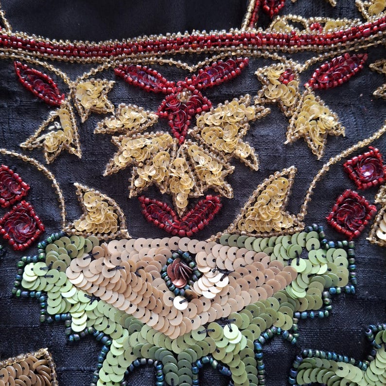 Puede incluir: Primer plano de un textil con intrincado bordado de cuentas y lentejuelas. El dise&ntilde;o presenta lentejuelas y cuentas doradas, rojas y verdes dispuestas en patrones florales y geom&eacute;tricos sobre un fondo oscuro. El bordado es detallado y ornamentado.