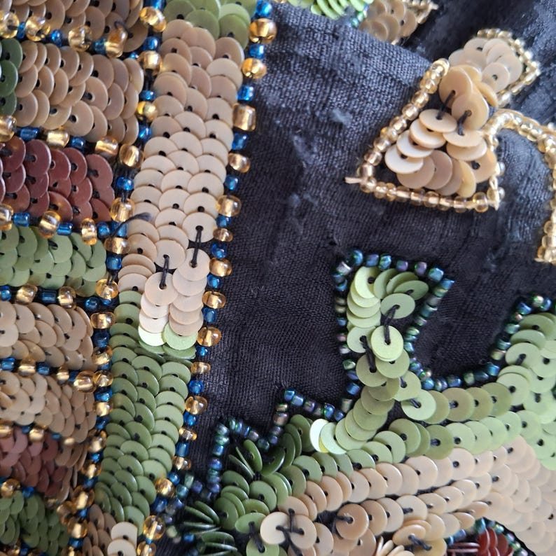 Puede incluir: Primer plano de una tela negra adornada con lentejuelas y abalorios. El dise&ntilde;o presenta un patr&oacute;n floral con lentejuelas verdes, marrones y beige, y abalorios azules y dorados. Las lentejuelas redondas crean una superficie texturizada.
