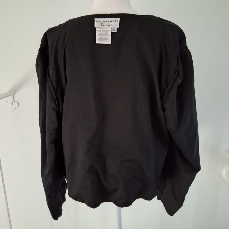 Puede incluir: Top negro de manga larga con cuello redondo. La parte superior tiene una etiqueta que dice "Adrianna Papell Evening" y es talla XL. La prenda est&aacute; hecha de una tela negra lisa y suave.