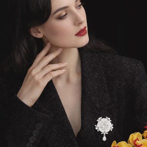 May include: A woman wearing a black blazer with a white floral brooch and a dangling pearl. The brooch is the focal point, set against the dark fabric. Yellow orchids add a pop of color.