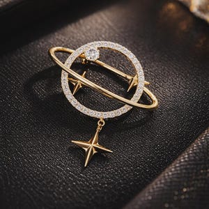 May include: Gold-toned brooch in a celestial design. The brooch features a ring with embedded crystals, a dangling star, and a central crystal. The brooch is set against a dark background.