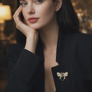 May include: A gold-toned bee brooch with a green center stone and white accents is pinned to a black blazer. The brooch is the focal point, set against the dark fabric. The image is a portrait of a person.