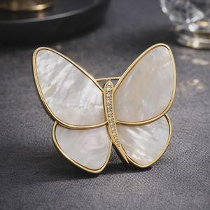 May include: A gold butterfly brooch featuring iridescent white wings. The wings are framed in gold, and the body is embellished with tiny, sparkling crystals. This elegant accessory is ideal for adding a touch of nature-inspired glamour.