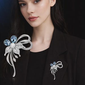 May include: A silver-toned flower brooch with blue gemstone accents and a central pearl. The brooch features a bow design and is adorned with clear crystals. A smaller version of the brooch is also visible.