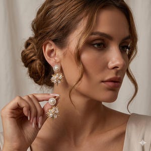 May include: Gold-toned, dangling earrings with a pearl stud and a starburst design. The earrings feature a large, round pearl at the top and a cluster of smaller crystals and gold accents below. The earrings are designed to be worn on the ear.
