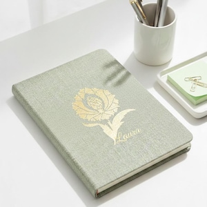 May include: A sage green notebook featuring a gold floral design and the name "Laura". The notebook is on a white surface, with pens in a white cup and a small tray with sticky notes and paper clips.