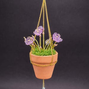 May include: A hanging terracotta pot with a gold chain and clasp. The pot contains green moss and wire flowers in shades of purple and gold. A decorative chain with beads hangs from the bottom of the pot.
