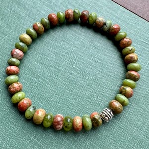 May include: A beaded bracelet featuring a variety of natural stone beads in shades of green, red, and brown. The beads are oval-shaped and strung together with a silver-colored metal accent bead. The bracelet is displayed on a green surface.