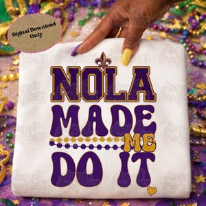 May include: White t-shirt with the text "NOLA MADE ME DO IT" in purple and gold lettering. The design includes a fleur-de-lis and a string of beads, evoking a festive theme. A digital download only.