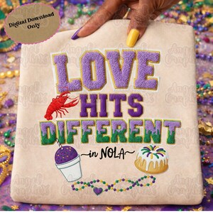 May include: A beige t-shirt with the text "LOVE HITS DIFFERENT in NOLA" in purple, green, and white. The design includes a red crawfish, a purple snow cone, and a king cake. A hand with purple and yellow nails is holding the shirt.