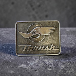 May include: A rectangular, antique-style metal belt buckle with the word "Thrush" in a stylized font. The buckle features a graphic of a bird-like creature with a circular background.