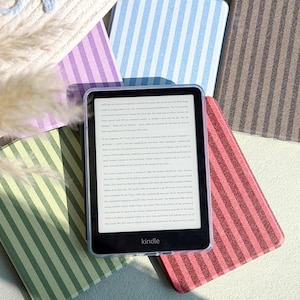 Puede incluir: Un lector de libros electrónicos Kindle con una funda transparente, rodeado de varias fundas Kindle a rayas en colores pastel. Las fundas son de color azul claro, rosa, verde, morado y marrón. La pantalla del Kindle muestra texto y la marca "Kindle" es visible.