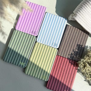 May include: A collection of rectangular, striped cardholders in various pastel colors, including lavender, blue, green, and pink. Each cardholder features a different set of initials in the corner. The cardholders are arranged on a light-colored surface.