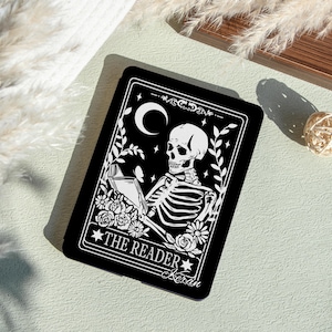 May include: A black rectangular card featuring a white illustration of a skeleton reading a book under a crescent moon. The card is decorated with floral and star motifs, with the text "THE READER" and the name "Kieran".