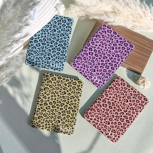 Puede incluir: Cuatro estuches rectangulares con estampados de leopardo en varios colores: azul claro, morado, beige y rosa. Cada estuche tiene una inicial diferente en la esquina inferior. Los estuches están colocados sobre una superficie azul claro.
