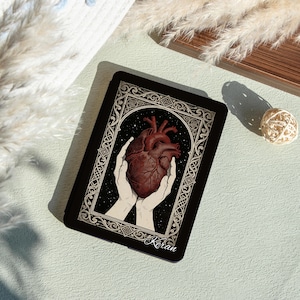 Könnte beinhalten: Schwarze Tablet-Hülle mit einer detaillierten Illustration eines menschlichen Herzens, das in zwei Händen gehalten wird, vor einem Sternenhintergrund. Das Design ist von einem verzierten Rahmen umgeben. Der Name "Kiran" ist unten aufgedruckt. Die Hülle liegt auf einer hellgrünen Oberfläche.
