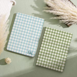 May include: Two rectangular tablet or e-reader cases featuring a gingham check pattern. One case is light blue and white with the letters SK printed in the corner, while the other is sage green and white with the letters TY printed in the corner.