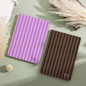 May include: Two rectangular tablet cases with vertical stripes. One case is light and dark purple with the letters SK in the bottom corner. The other case is dark brown and black with the letters TY in the bottom corner. Both have a smooth finish.