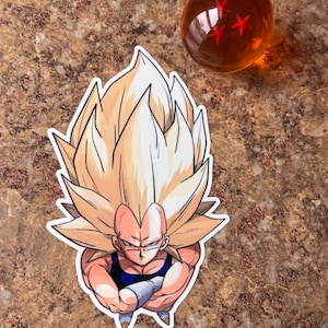 Puede incluir: Una pegatina con un personaje de pelo rubio puntiagudo, un atuendo azul y negro y una expresión decidida. Junto a ella hay una esfera naranja transparente con tres estrellas rojas en su interior. La pegatina está sobre una superficie marrón.