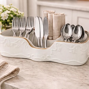 May include: A white ceramic utensil holder with a gold trim, filled with forks, knives, spoons, and rolled napkins. The holder has a decorative embossed design. A bottle of oil and a bouquet of white flowers are in the background.