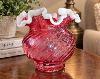 May include: A cranberry red glass vase featuring a wide, ruffled rim with a thick, opaque white glass edge. The body of the vase has a distinct spiral ribbed texture throughout, creating a swirling pattern in the translucent red glass material.