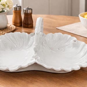May include: White ceramic serving dish with a handle, featuring a leaf-like design. The dish is divided into two sections and sits on a wooden table. Other items on the table include a bowl of fruit and a vase of flowers.