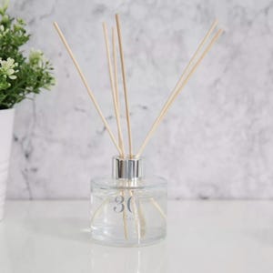 Op de afbeelding: Een heldere glazen diffuserfles met een zilveren kraag en houten rietjes. De fles heeft het nummer "30" op de voorkant gedrukt. De diffuser staat op een wit oppervlak met een marmeren achtergrond.