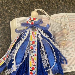 May include: A blue and white tassel with ribbons, lace, and a wooden bead. The tassel has ribbons that say "Teach, Inspire, Love." A heart-shaped charm and two pearl beads are attached to the tassel. The tassel is resting on an open book.