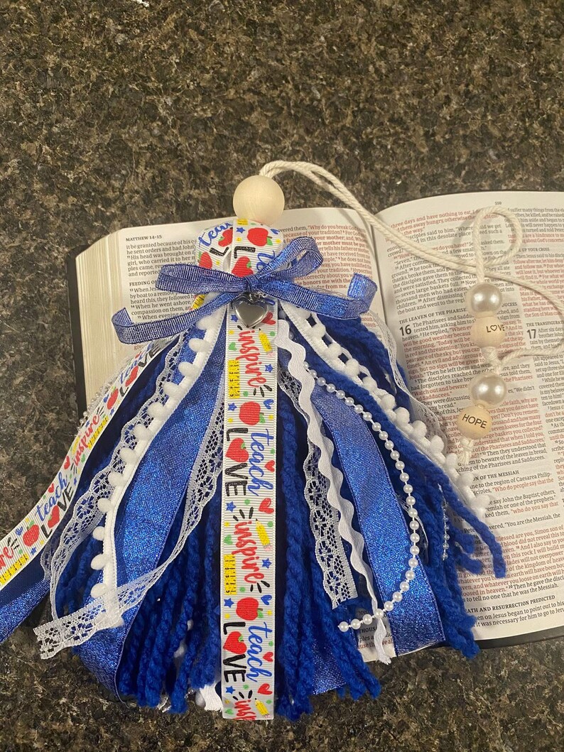 May include: A blue and white tassel with ribbons, lace, and a wooden bead. The tassel has ribbons that say "Teach, Inspire, Love." A heart-shaped charm and two pearl beads are attached to the tassel. The tassel is resting on an open book.