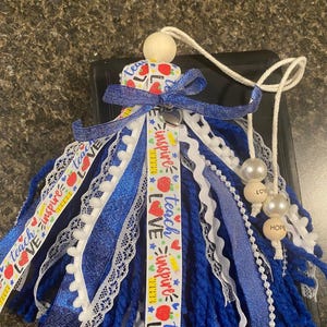 May include: A handmade tassel ornament with blue yarn, white lace, and ribbons. The ribbons feature the words "Teach, Inspire, Love" and apple graphics. The ornament includes wooden beads and pearls, with the words "Love" and "Hope".