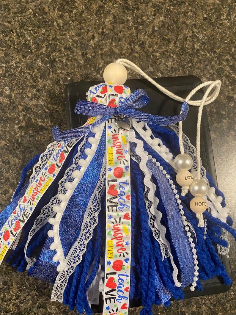 May include: A handmade tassel ornament with blue yarn, white lace, and ribbons. The ribbons feature the words "Teach, Inspire, Love" and apple graphics. The ornament includes wooden beads and pearls, with the words "Love" and "Hope".