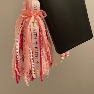 May include: A pink and white tassel bookmark with the word "FAITH" printed on a ribbon, accented with a gold cross charm. The tassel is attached to a wooden bead and is next to a black book.