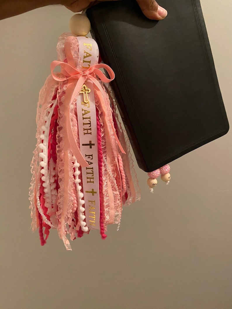 May include: A pink and white tassel bookmark with the word "FAITH" printed on a ribbon, accented with a gold cross charm. The tassel is attached to a wooden bead and is next to a black book.