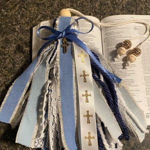 May include: A handmade angel ornament with blue and white ribbons, lace, and a wooden bead head. A blue ribbon with a gold cross is tied around the head. A white ribbon with gold crosses is also present. The ornament is displayed on an open book.