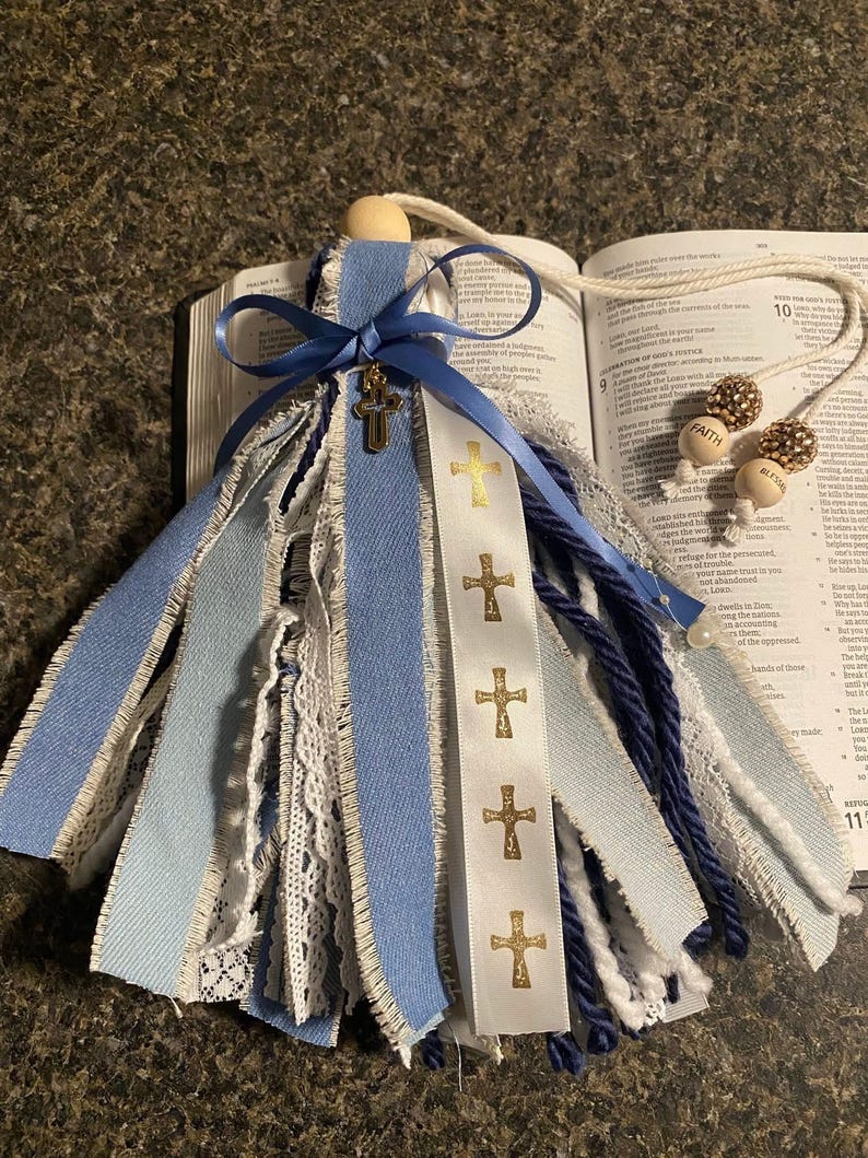 May include: A handmade angel ornament with blue and white ribbons, lace, and a wooden bead head. A blue ribbon with a gold cross is tied around the head. A white ribbon with gold crosses is also present. The ornament is displayed on an open book.