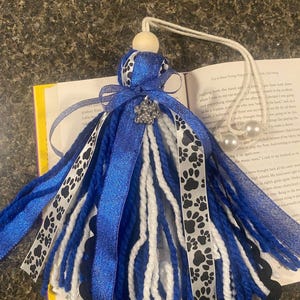 May include: A blue and white tassel with paw print ribbon and a decorative jewel. The tassel is made of blue and white yarn and has a wooden bead at the top. The tassel is approximately 8 inches long.