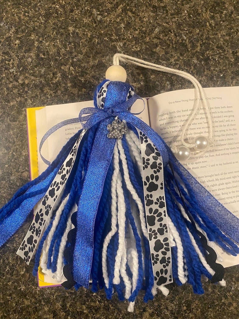 May include: A blue and white tassel with paw print ribbon and a decorative jewel. The tassel is made of blue and white yarn and has a wooden bead at the top. The tassel is approximately 8 inches long.