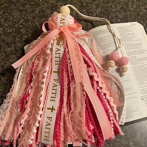 May include: A pink and white tassel ornament with the word "FAITH" printed on a ribbon, accented with a cross. The ornament has a wooden bead at the top and a string for hanging. Two beaded ornaments with the words "BLESSED" and "LOVE" are attached.