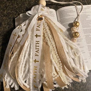 May include: A handmade tassel with white and gold ribbons, lace, and yarn. The tassel features the word "FAITH" printed on a white ribbon with gold crosses. Wooden beads with the words "LOVE" and "BLESS" are attached to a string.