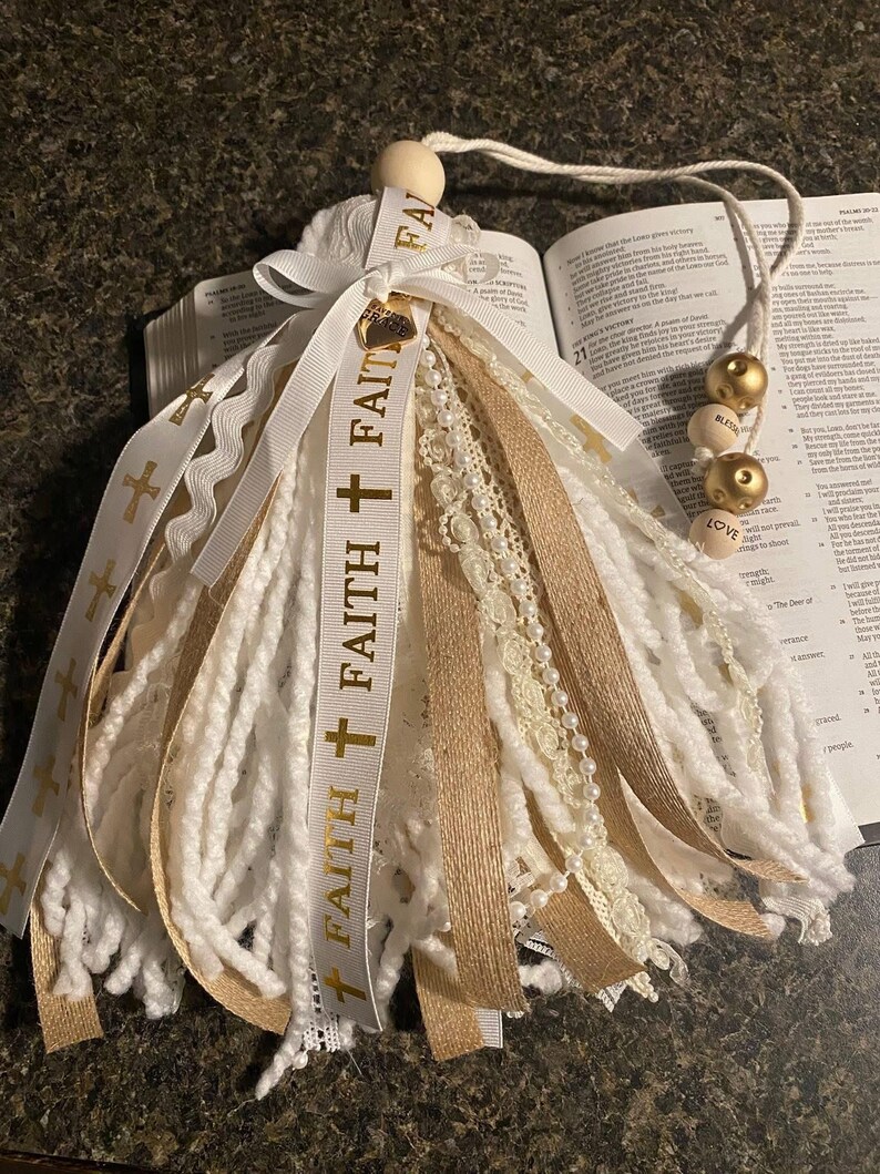 May include: A handmade tassel with white and gold ribbons, lace, and yarn. The tassel features the word "FAITH" printed on a white ribbon with gold crosses. Wooden beads with the words "LOVE" and "BLESS" are attached to a string.