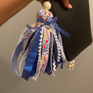 May include: A blue, white, and red tassel with the words "Teach, Inspire, Love" on a ribbon, a wooden bead, and a silver heart charm. The tassel is attached to a black tablet.