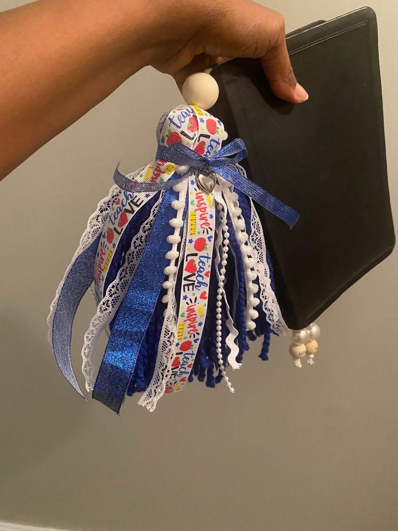 May include: A blue, white, and red tassel with the words "Teach, Inspire, Love" on a ribbon, a wooden bead, and a silver heart charm. The tassel is attached to a black tablet.