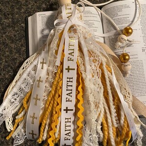 May include: A decorative tassel featuring white, yellow, and cream-colored yarn and lace. The tassel includes a wooden bead and a ribbon printed with the word "FAITH" and cross symbols. Gold beads and a cord are attached.