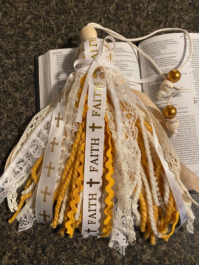 May include: A decorative tassel featuring white, yellow, and cream-colored yarn and lace. The tassel includes a wooden bead and a ribbon printed with the word "FAITH" and cross symbols. Gold beads and a cord are attached.