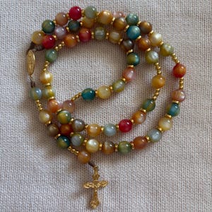 May include: A rosary featuring multicolored, translucent, polished beads in shades of red, blue, green, and yellow, separated by small gold-toned metal spacers. The rosary includes a gold-toned metal crucifix and a small oval religious medal.
