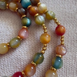 May include: A multi-strand necklace featuring smooth, round, multicolored beads in shades of blue, green, yellow, orange, and pink. Each large bead is separated by small, polished gold-tone metal spacer beads, creating a vibrant, reflective pattern.