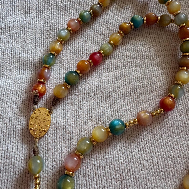 May include: A rosary featuring multicolored translucent beads in shades of green, yellow, orange, red, and blue, separated by small gold-toned metal spacers. An oval gold-toned metal centerpiece displays an embossed cross and the letter M.