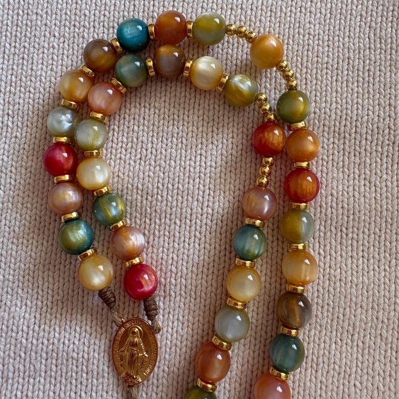 May include: A rosary featuring multicolored, polished, translucent beads in shades of blue, green, yellow, orange, and red. Each bead is separated by small gold-toned metal spacers. A gold-toned oval medal depicting a figure is attached.