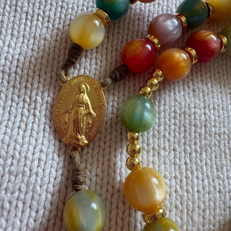 May include: A rosary featuring multicolored, polished, spherical beads in shades of yellow, green, red, and purple, separated by small gold-toned metal spacers. A gold-toned oval medal depicting a figure in relief is attached to the brown cord.