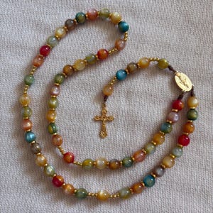 May include: A rosary featuring multicolored, translucent, polished stone beads in shades of red, blue, green, and yellow, separated by small gold-toned metal spacers. The piece includes a gold-toned metal centerpiece and a detailed gold-toned crucifix.